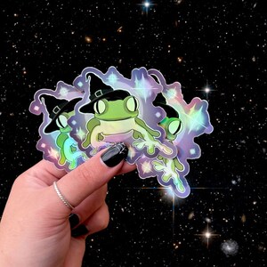 Puede incluir: Tres pegatinas holográficas con una rana verde que lleva un sombrero de bruja y está rodeada de estrellas. Las pegatinas son iridiscentes y tienen un tema mágico.