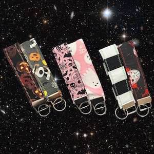 May include: Five Halloween-themed keychains with different designs. The keychains are made of fabric and have a variety of colors, including black, white, red, green, and pink. The keychains feature designs such as pumpkins, ghosts, bats, and a skull.