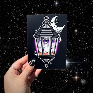 Puede incluir: Una ilustración en blanco y negro de una linterna con tres fantasmas en su interior. La linterna brilla en púrpura y tiene una luna creciente y murciélagos por encima. Hay calabazas y telarañas dentro de la linterna.
