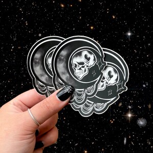 May include: A stack of black and white stickers featuring a skull inside a crescent moon design. The stickers have a decorative border and star accents. The background is a starry night sky.