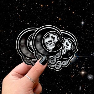 May include: Black and white stickers featuring a skull inside a crescent moon design. The stickers have a detailed, gothic aesthetic with a celestial background. The stickers are held in a hand with a black painted nail.