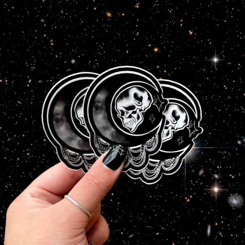May include: Black and white stickers featuring a skull inside a crescent moon design. The stickers have a detailed, gothic aesthetic with a celestial background. The stickers are held in a hand with a black painted nail.