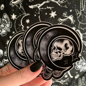 May include: A stack of black and white stickers featuring a skull inside a crescent moon design. The stickers have a decorative lace border and are held against a black background with white celestial and occult-themed patterns.