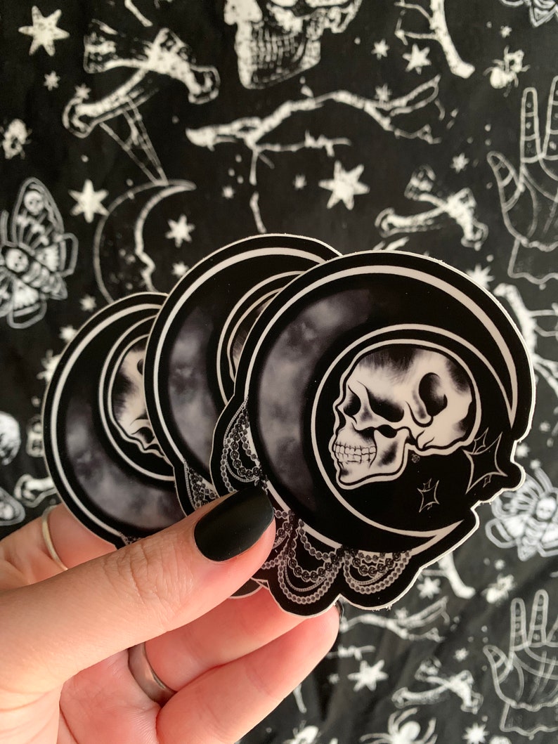 May include: A stack of black and white stickers featuring a skull inside a crescent moon design. The stickers have a decorative lace border and are held against a black background with white celestial and occult-themed patterns.
