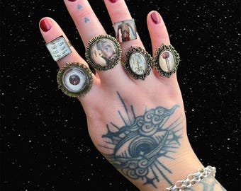 May include: A hand wearing six rings with different designs. The rings feature images of an eye, a skeleton, a flower, a human anatomy diagram, and a portrait. The hand has a tattoo of an eye with clouds on the back.