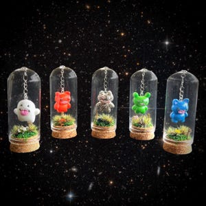 Op de afbeelding: Vijf miniatuur terrariums met cartoonfiguren in glazen koepels. Elke koepel heeft een kurken basis en een klein stukje groen. De figuren zijn wit, rood, met patronen, groen en blauw, opgehangen aan kettingen tegen een sterrenachtergrond.