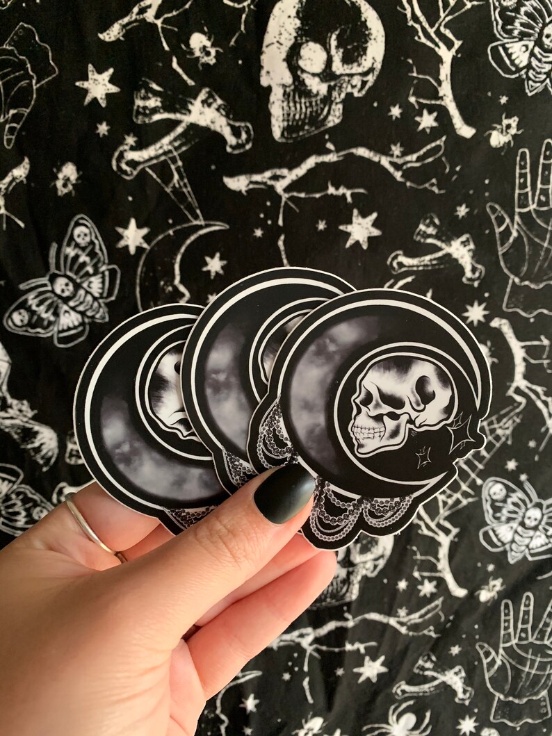 May include: A stack of black and white stickers featuring a skull inside a crescent moon design. The stickers are held against a black background with a repeating pattern of skulls, moths, and stars.