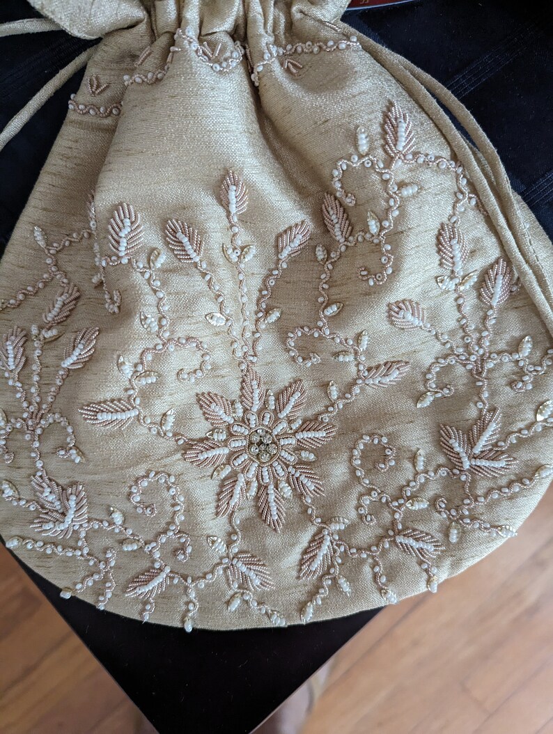 May include: A beige drawstring bag with intricate embroidery featuring a floral design with white and pink beads.