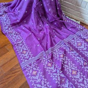 May include: A vibrant purple textile with intricate white and gold geometric patterns along the borders and scattered polka dots. The fabric is draped, showcasing its texture and design. The textile appears to be a saree or similar garment.
