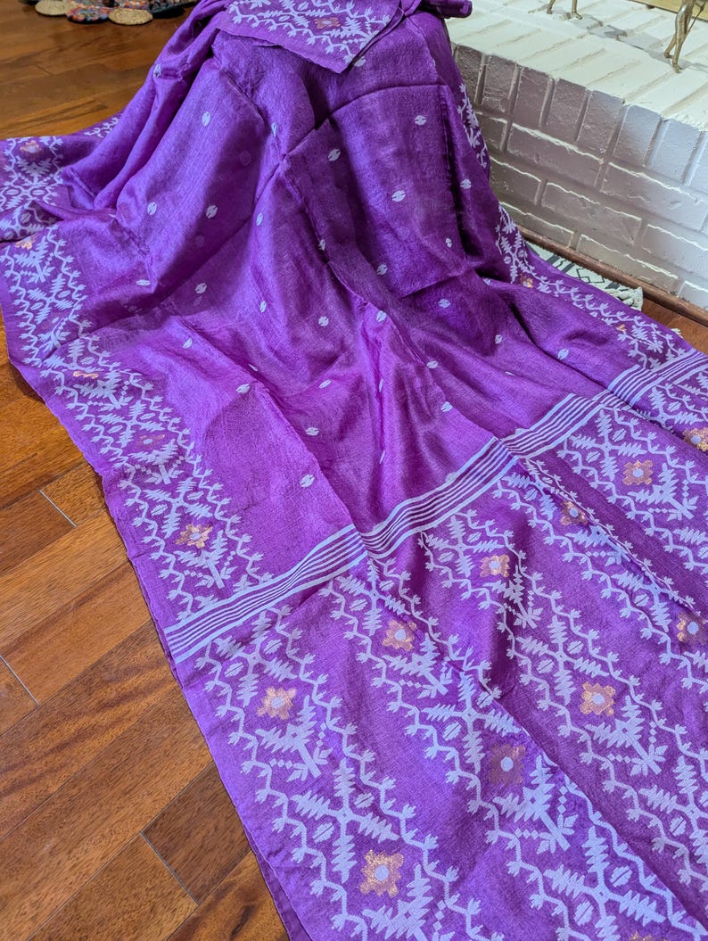May include: A vibrant purple textile with intricate white and gold geometric patterns along the borders and scattered polka dots. The fabric is draped, showcasing its texture and design. The textile appears to be a saree or similar garment.