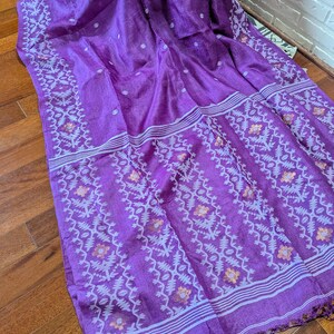 May include: A vibrant purple textile with intricate white and orange floral patterns along the borders and stripes. The fabric is draped, showcasing its texture and design. The textile has a fringed edge.