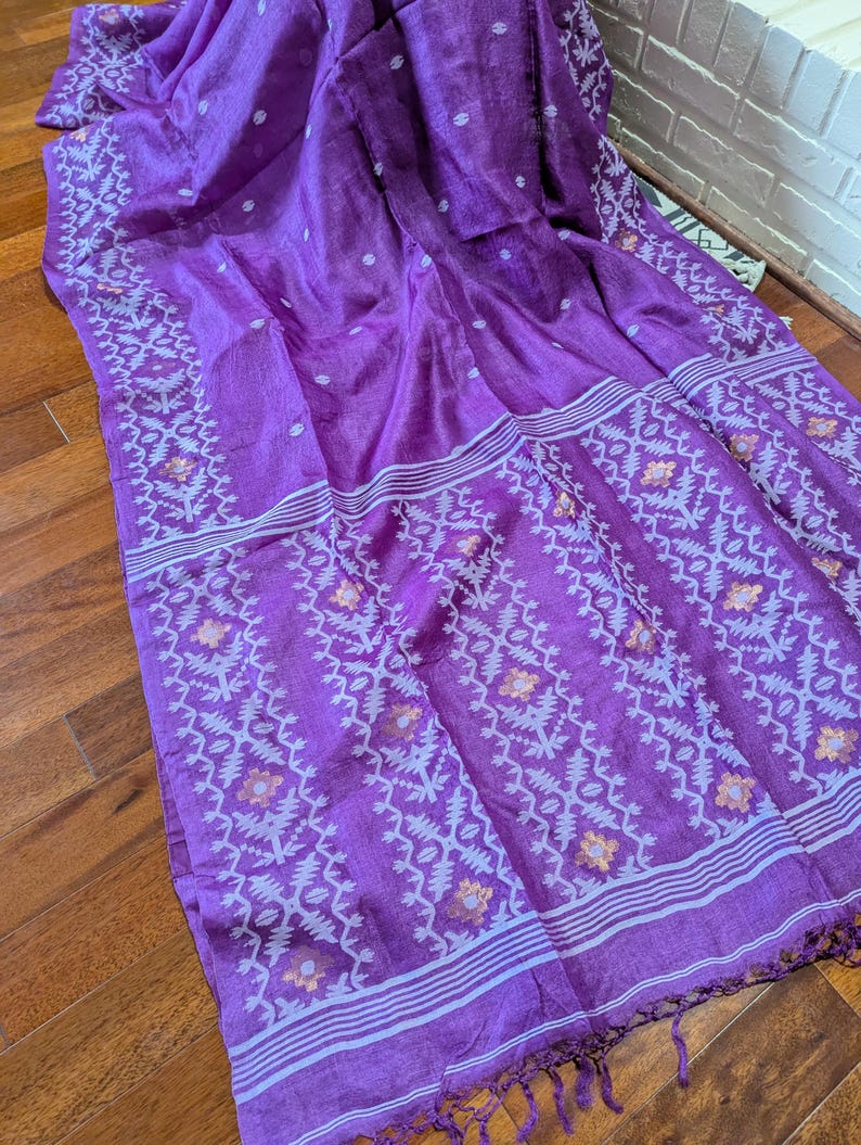 May include: A vibrant purple textile with intricate white and orange floral patterns along the borders and stripes. The fabric is draped, showcasing its texture and design. The textile has a fringed edge.