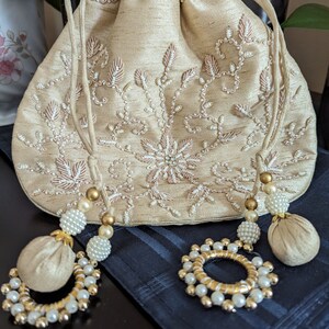 May include: A beige drawstring bag with a floral design embroidered with white beads. The bag has two tassels with gold beads and white pearls.