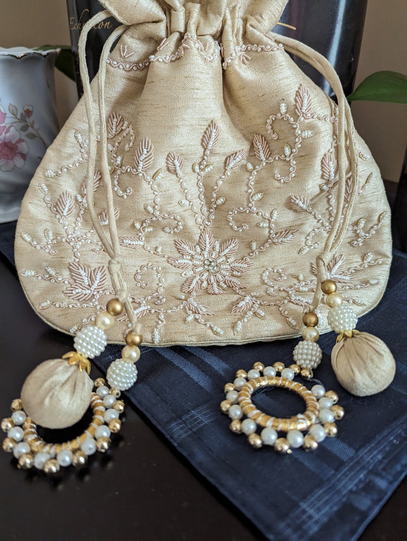 May include: A beige drawstring bag with a floral design embroidered with white beads. The bag has two tassels with gold beads and white pearls.