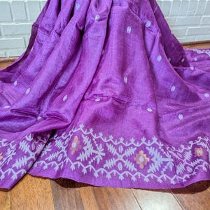 May include: A vibrant purple textile with a decorative border. The fabric is draped, showcasing its texture and design. The border features a repeating geometric pattern in white and gold. The textile is likely a saree or similar garment.