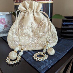 May include: A beige drawstring bag with white embroidery and pearl accents. The bag has two tassels with gold beads and pearls.