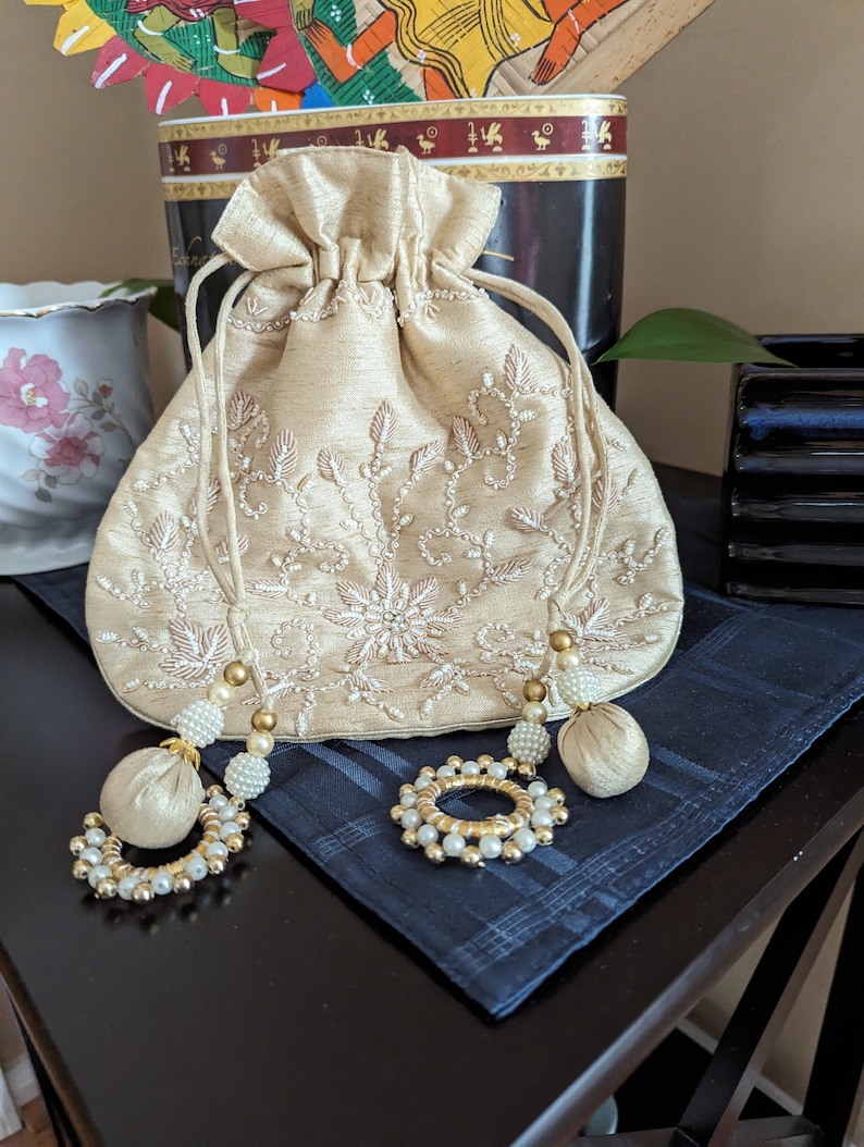 May include: A beige drawstring bag with white embroidery and pearl accents. The bag has two tassels with gold beads and pearls.
