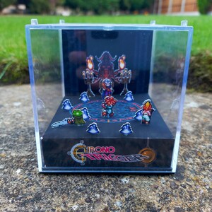 Könnte beinhalten: Ein durchsichtiger Würfel-Präsentationskoffer aus Kunststoff mit schwarzem Hintergrund, der ein 3D-Diorama einer Videospielszene zeigt. Die Szene zeigt Charaktere aus dem Spiel Chrono Trigger, darunter eine rothaarige Figur mit einem Schwert, eine grüne froschartige Figur und eine rothaarige Figur mit einem Stab. Der Hintergrund zeigt eine große, bedrohliche Gestalt mit Hörnern und einem roten Umhang. Die Basis des Präsentationskoffers trägt das Logo des Spiels, "Chrono Trigger".
