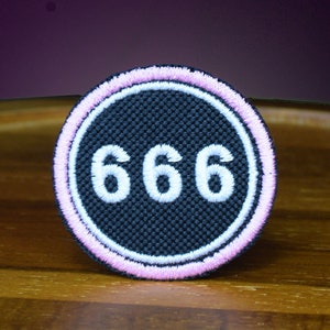 May include: A black and white embroidered patch with the number 666 in white on a black background. The patch has a pink border.