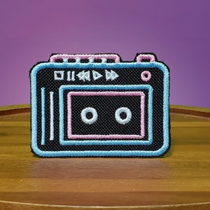 Puede incluir: Un parche negro en forma de reproductor de casetes con contornos azules y rosas. El parche tiene un botón de rebobinado blanco, un botón de avance rápido blanco y un botón de reproducción blanco. La cinta de casete es negra con dos círculos blancos.