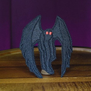 Puede incluir: Parche bordado negro de una criatura con alas grandes y ojos rojos. La criatura está de pie sobre una superficie de madera.