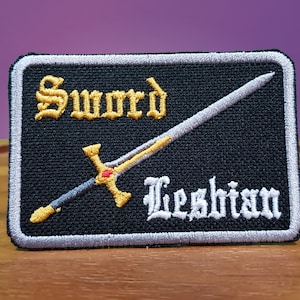 Puede incluir: Un parche bordado negro con un contorno plateado. El parche presenta una espada dorada con una joya roja y el texto "Sword Lesbian" en blanco.