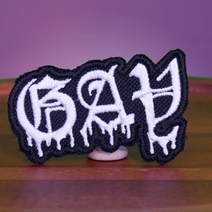May include: Black and white embroidered patch with the word "GAY" in dripping white letters.