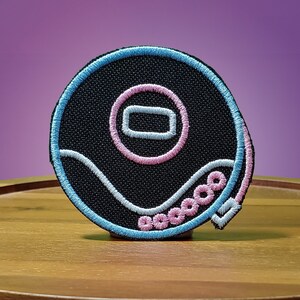 Puede incluir: Un parche bordado negro con un contorno azul y rosa. El parche presenta un diseño estilizado de un disco con una línea ondulada y un círculo en el centro.