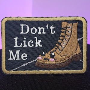 Peut inclure: Un écusson brodé noir et or avec une botte marron et le texte "Don't Lick Me".