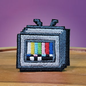 May include: A gray embroidered patch of a retro television with a pixelated design and a colorful test pattern on the screen.