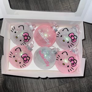 May include: Six glittery Christmas baubles in a white box, each featuring Hello Kitty's face. Some are pink, some white, and some have "Hello Kitty" written on them. The baubles have pink bows and whiskers.