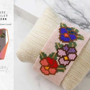 Pode incluir: Um bracelete de contas rosa com um design floral. O bracelete apresenta três flores em tons de vermelho, rosa, azul e amarelo. O bracelete está sobre um fundo de tecido branco.