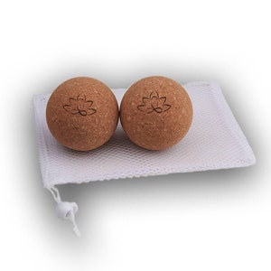 May include: Two brown cork massage balls with a lotus flower design, resting on a white mesh bag. The balls are spherical and appear to be made of natural cork material. The bag has a drawstring closure.