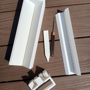 Könnte beinhalten: Ein Set aus weißen Kunststoffkomponenten, bestehend aus einer langen rechteckigen Rinne, einem kleineren spitzen Marker, einem gekerbten Basisteil und einem weißen Lineal mit Markierungen von 0,5 bis 9,0 cm, sowie einem dünnen Metallwerkzeug mit dunklem Holzgriff.