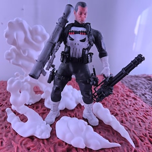 May include: Action figure of a character in a black tactical suit with a white skull emblem on the chest. The figure is holding a large weapon and a machine gun. White smoke-like shapes are around the figure.