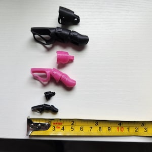 Puede incluir: Surtido de piezas de mano de robot impresas en 3D en negro y rosa. Las piezas est&aacute;n dispuestas sobre una superficie blanca con una cinta m&eacute;trica amarilla.