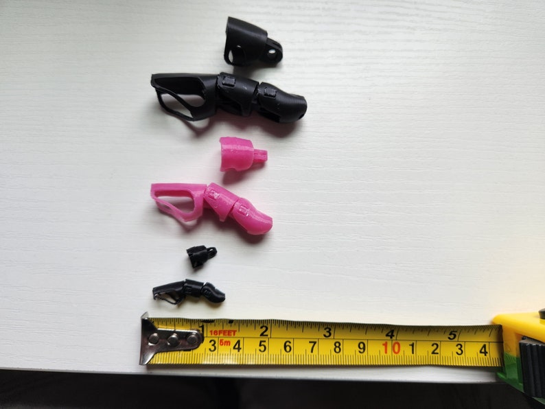 Puede incluir: Surtido de piezas de mano de robot impresas en 3D en negro y rosa. Las piezas est&aacute;n dispuestas sobre una superficie blanca con una cinta m&eacute;trica amarilla.