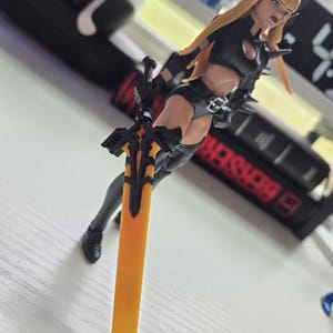 May include: A figurine of a female warrior with long blonde hair, wearing a black outfit and holding a large orange and black sword. The figure has a stern expression and is posed dynamically. The background is blurred.