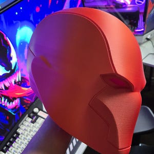 May include: A bright red, textured helmet, likely for cosplay or costume use. The helmet has a smooth, rounded design with defined facial features and a textured surface. The overall shape suggests a superhero or comic book theme.