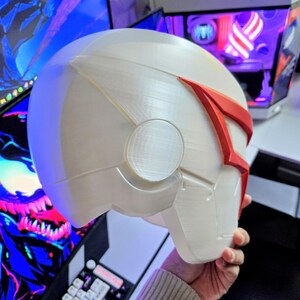 May include: A white and red helmet with a futuristic design. The helmet has a smooth, rounded shape with angular details and a red accent across the top. The helmet is held in a person's hand.