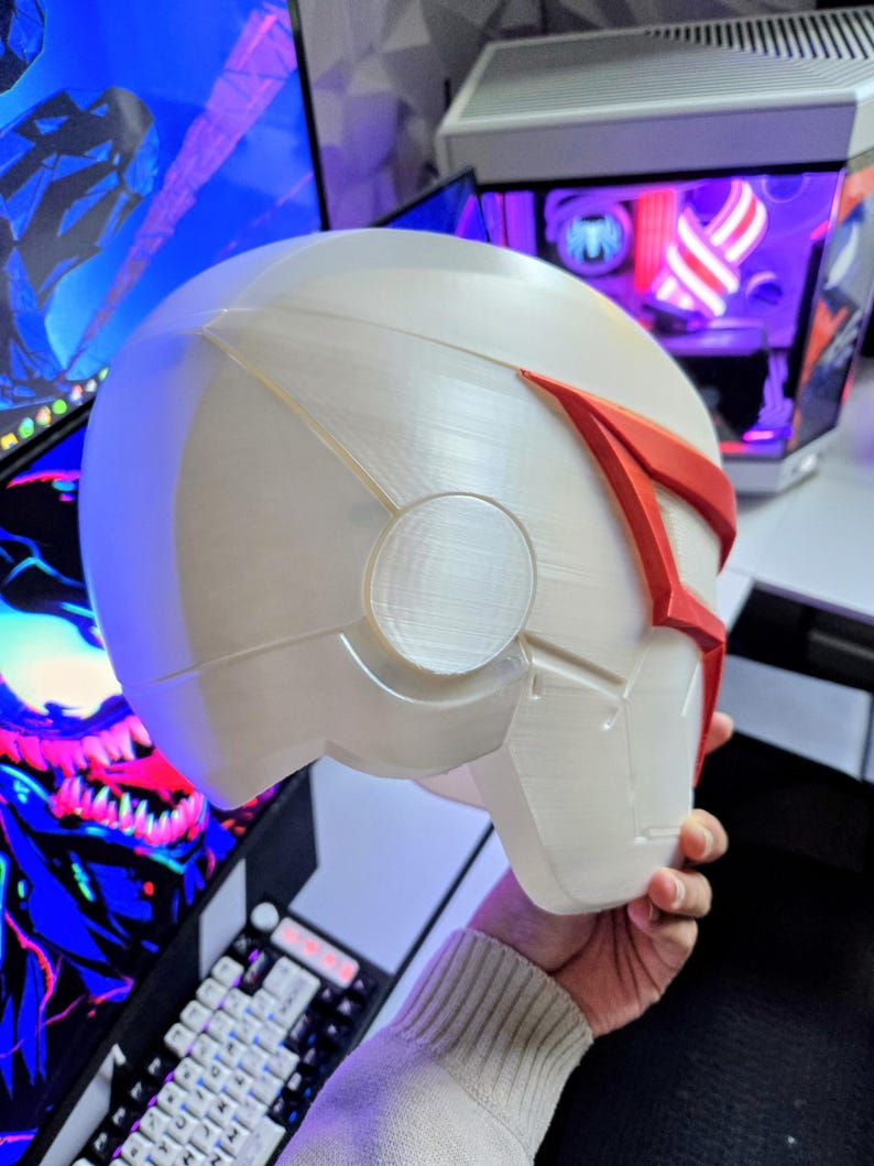 May include: A white and red helmet with a futuristic design. The helmet has a smooth, rounded shape with angular details and a red accent across the top. The helmet is held in a person's hand.