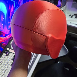 May include: A red, textured helmet with a distinctive design, possibly for cosplay or a costume. The helmet has a smooth, rounded top and a textured surface. The design includes a face covering. The helmet is held in front of a computer setup.