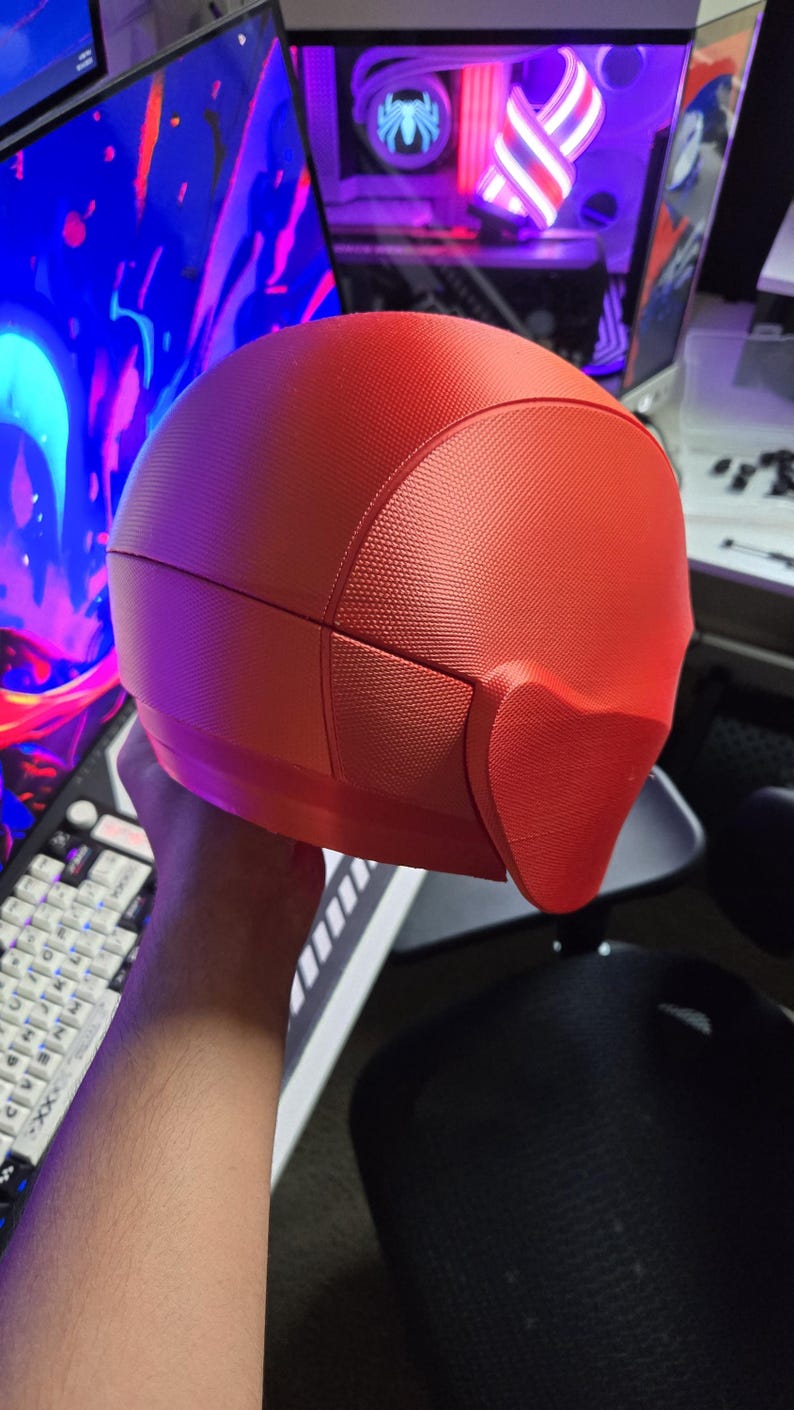 May include: A red, textured helmet with a distinctive design, possibly for cosplay or a costume. The helmet has a smooth, rounded top and a textured surface. The design includes a face covering. The helmet is held in front of a computer setup.