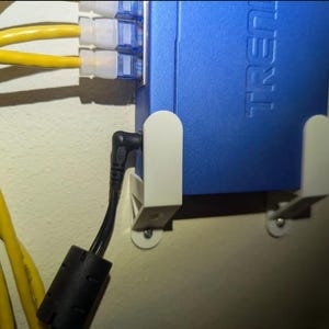Puede incluir: Un router de red azul montado en una pared blanca con un cable de alimentación negro enchufado. El router tiene la palabra "TRENDnet" impresa en él.