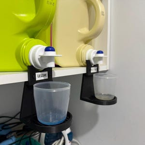 Può includere: Sistema di erogazione del detersivo per bucato con due contenitori. Uno è verde lime, l'altro beige. Ognuno ha un beccuccio bianco con un tappo blu. Le staffe nere sostengono bicchieri dosatori trasparenti. La parola "LESSIVE" è su un'etichetta.