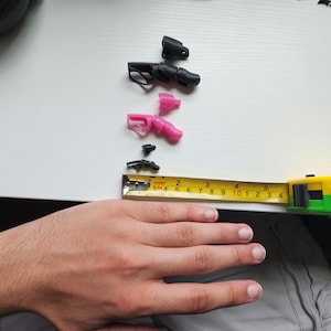 Puede incluir: Primer plano de una mano junto a una cinta m&eacute;trica y varias piezas peque&ntilde;as de pl&aacute;stico negro y rosa. La cinta m&eacute;trica est&aacute; marcada en cent&iacute;metros y lee 25,4 cm.