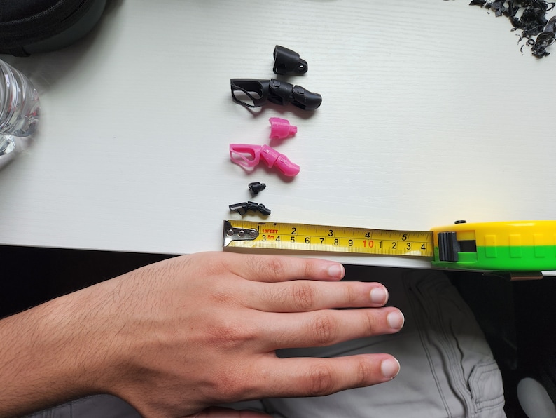 Puede incluir: Primer plano de una mano junto a una cinta m&eacute;trica y varias piezas peque&ntilde;as de pl&aacute;stico negro y rosa. La cinta m&eacute;trica est&aacute; marcada en cent&iacute;metros y lee 25,4 cm.