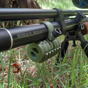 May include: A black air rifle with a scope and a green grenade-shaped accessory rests in long green grass. The rifle has a wooden stock and is supported by a black bipod. The text "MAXIMUM FILL PRESSURE OF 30" is visible on the rifle.