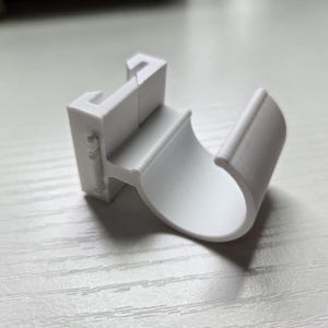 May include: A white plastic cable clip with a curved design. The clip has a hook-like feature on one end and a rounded section to hold a cable. The clip is designed to secure and organize cables.
