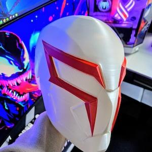May include: A white and red superhero mask with a geometric design. The mask has red accents around the eyes and edges, with a textured surface. The mask is likely 3D printed and is a cosplay or costume item.